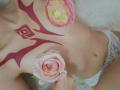 AmeliaMaiv's bare chest with red tribal tattoos, holding pink roses covering her tits, wearing white lace panties, lying on a fluffy white surface.