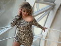 CandyLadyZ with curly blonde hair, wearing a leopard print dress, showing her large boobs and smiling, leaning on a white railing, in a bright room with glass windows.