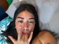 SharonKlart with dark hair, brown eyes, red nails, and pink lipstick, sucking on her finger, lying on a bed, grey pillow, and blue-green patterned sheets.