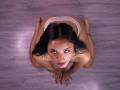 ArisSuarez, a woman with black hair and fair skin, kneels on a wooden floor, looking up with intense eyes. She wears white lace panties, her hands pressed against the floor.