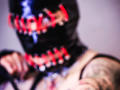 AmySpider wearing a black mask with red and white stitches, hands near mouth, tattooed arm visible, blurred face, purple-gray background, suggestive, edgy, intense.