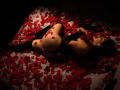 SophiaXoxo lying on a bed covered in red rose petals, wearing black thigh-high stockings and gloves, with her ass and back exposed, in dim, sensual lighting.