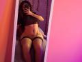 Sofia Jackson, met krullend haar, een zwarte doorschijnende top, netpanty's en een tatoeage op haar rechterheup, maakt een spiegelzelfportret waarop haar borsten en vagina te zien zijn..