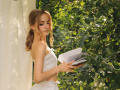 EvaDaly with small tits, wearing a white, strapless, ruffled dress, standing by green leaves, holding a book, sunlight filtering through, in a peaceful outdoor setting.