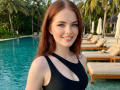 BryannaCrecelius with red hair, smiling, wearing a black one-piece swimsuit with a cutout, showing her small tits, by a blue pool with wooden loungers and palm trees.