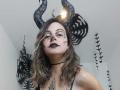 ArianaBlonde with black glitter horns, dark makeup, and black lipstick, wearing a choker and glasses, showing tattoos on chest and neck, with wavy brown hair.