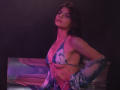 StellaLewin with pink-tinged skin, wearing a tie-dye bikini top and jacket, exposing her tits, with a smoky purple and pink light, leaning against a reflective surface.