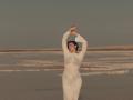 OliviaCluff in a white, long-sleeved dress, arms raised, standing on a beach, eyes closed, with soft light and calm water in the background.
