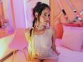 MelisaDaltonn, femme asiatique aux cheveux tressés, portant une robe blanche transparente, montrant ses seins et ses parties intimes, un sac jaune scintillant, assise sur une chaise rose, éclairage rose et violet..