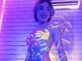 GabySmiith has neon paint on her tits, ass, and chest, with colorful handprints. She's smiling, standing in a purple-lit room with a neon sign behind her.