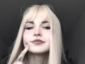 EmelyDilsaver with blonde hair, pinkish skin, and closed eyes, holding her chin with a finger, showing tattoo on finger, and soft brown lipstick, in a gray-lit room.