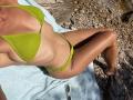 SandraSwweet lying on a towel, wearing a green ribbed bikini, sunlit, tan skin, small tits, flat stomach, green thong, sunglasses near her butt, rocky ground, legs crossed.