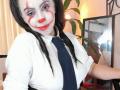KimFire with painted white face, red nose, and red smile, wearing a white shirt and black tie, has black braids, and is holding a mirror, showing her tits.