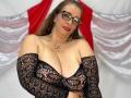 Miranda Delacroix mit großen Brüsten, trägt schwarze Spitzendessous, eine Brille und roten Lippenstift, hat braunes Haar und eine Halskette, lächelt, im Hintergrund befindet sich ein rot-weißes Tuch..