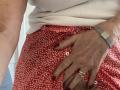 Bourgeoisex's hand with red nail polish and gold ring rests on her red floral skirt, covering her pussy. She wears a white top and a gray watch.