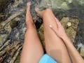 SandraSwweet's tan legs with white nails, sitting in clear water, her hand on her thigh, wearing blue shorts, with rocky bottom visible, water splashing around her feet.