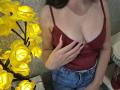 Rouzi in a red tank top, holding her tits with one hand, wearing blue jeans, yellow rose lamps in the background, standing indoors.