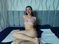 AbbyHillsI with small tits in pink floral bra, sitting on bed, legs crossed, smiling, wearing pink panties, green hair, pale skin, grey headboard, blue and white bed.