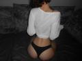 ErynAttridge kneels on a dark bed, back to the camera, wearing a white, long-sleeve crop top and black thong, exposing her rounded, bare buttocks. Dark, textured pillows in background.