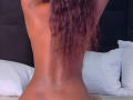 Shaaron's back, nude, with curly pinkish-brown hair, arms raised, white pillows in the background, her bare ass slightly visible, on a bed.
