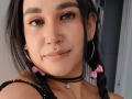 EdenIvyLm, avec ses cheveux noirs attachés en couettes, ses rubans roses, ses piercings au nez, son piercing au septum, ses boucles d'oreilles en forme de cerceaux, son collier noir, son collier avec un crâne et un maquillage discret, souriant doucement..