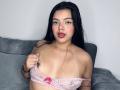 ViolettaRouse with long black hair, light brown skin, and red lipstick, pulling down a pink lace bra, exposing her small tits and visible tan lines, sitting on a gray couch.