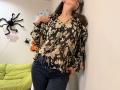 Tilliana, com cabelos castanhos longos, vestindo uma blusa marrom com estampas florais, calças jeans escuras e óculos, apoiada em uma parede, com as mãos nos bolsos, cabeça ligeiramente inclinada, olhando para cima..