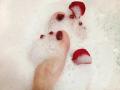VanesaMoka's foot with red-painted toenails, partially submerged in white foam, surrounded by red rose petals. Her toes are slightly curled, adding a sensual touch.