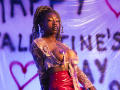 DaniaMarcell with dark skin, braided hair, wearing a sheer lace top with purple pasties over her big tits, red vinyl pants, and a tattoo on her lower abdomen.