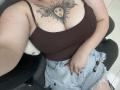 DiLaria with large tits, brown tank top, sun tattoo on chest, frayed denim shorts, red nails, holding right hand, sitting on black chair, visible arm, selfie.
