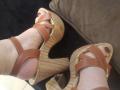 Close-up of SamanthaAntolin's feet in brown leather and wood platform sandals. Her toenails are painted dark brown. The sandals have criss-cross straps, and the wood platforms have a textured, woven pattern. Her skin is light, and the sandals are slightly worn. The right foot is slightly above the left, both pointing downward. The image focuses on the sandals and feet, with a brown cushion in the background. The texture of the wood platforms is prominent.