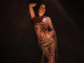 TriciaBarnes with bound tits and ass, wearing a rope skirt, leaning against dark wall, looking to the side, in dim light. Her hand rests on her hip, rope in front.