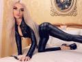 MademoiselleBella with long blonde hair, black leather outfit, and high heels, on a bed, on all fours, showing her ass, with a sexy, confident expression, holding a whip.