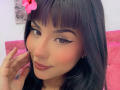 GraceClarke with dark hair, pink flower, and black eyeliner, has a nose piercing, glossy red lips, and a cute smile, touching her cheek. Pink headboard in background.