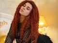 Niferttiti with long curly red hair, wearing a black fishnet top, leaning forward with eyes closed, pouty lips, and a nose ring. Her ass is partially covered by a black skirt.