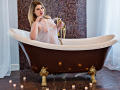Karolichee in a brown and gold clawfoot bathtub, holding a sheer lace top over her tits, with candles on the floor, water in the tub, and blonde hair.