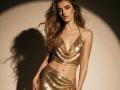 Feofaniia with long, wavy blonde hair, wearing a shiny, gold, sparkly halter top and skirt, showing her small, perky tits and slim, sexy ass, standing in shadow.
