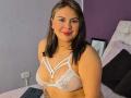 CamillaMoretti with medium tits in a white lace bra, smiling at the camera, brown hair, pink lipstick, nose ring, bed with black pillow, white wall plug in background.