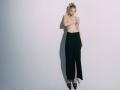 Topless EolantaMarina with blonde hair in a messy bun, wearing black high-waisted pants and black high heels, standing against a plain wall, covering her small tits.