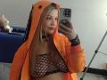 Melani Becker, met lang blond haar, draagt een oranje hooded jas, een zwarte nethemd en een choker, en maakt een spiegelzelfportret, waarbij haar borsten zichtbaar zijn, in een informele setting..