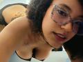 ScarlethVictoria with curly hair, glasses, and dark lipstick, bending over, showing her tits and black lace panties, tattoo on chest, necklace, smiling at camera.