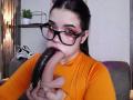 AryButty with dark hair and glasses, wearing an orange shirt, sucking a large, black, dildo-like object. Her eyes are heavily lined with red makeup.