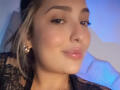BiancaRey's face, with glossy pink lips, thick eyebrows, and blonde hair in a black lace top, showing a slight smile, wearing gold earrings, in a soft blue light.