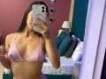 LaurentRay with medium tits in a pink bikini top, holding a phone in front of her face, standing in a bedroom with a bed in the background.