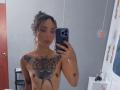 SilvanaXOwen with curly brown hair, topless, showing large butterfly tattoo on chest, small tits, holding phone in mirror selfie, gray wall, red door, and hanging light in background.