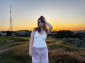 LeScharwath stands in a grassy field at sunset, wearing a white tank top with small studs and pink velvet pants. She has brown hair and a hand in it.