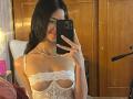 LiaBlom with long dark hair, smiling, wearing a lace white top showing her small tits, taking a mirror selfie with a black phone, pearl necklace. Bedroom setting.
