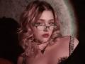 AdrianePittillo has pink curly hair, green glasses, and a black choker with a heart pendant. She's showing her tits in a low-cut top. Her face is slightly shadowed.