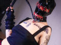AmySpider in black latex mask with red laces, holding a black whip, wearing a black tank top, exposing tattoos on chest and arms, with black fingerless gloves.