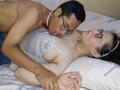 CoupleFantasticErotic man with short black hair and glasses, shirtless, licking the neck of CoupleFantasticErotic woman in white lace top, lying on bed with white pillows, holding hands. Sexual tension.